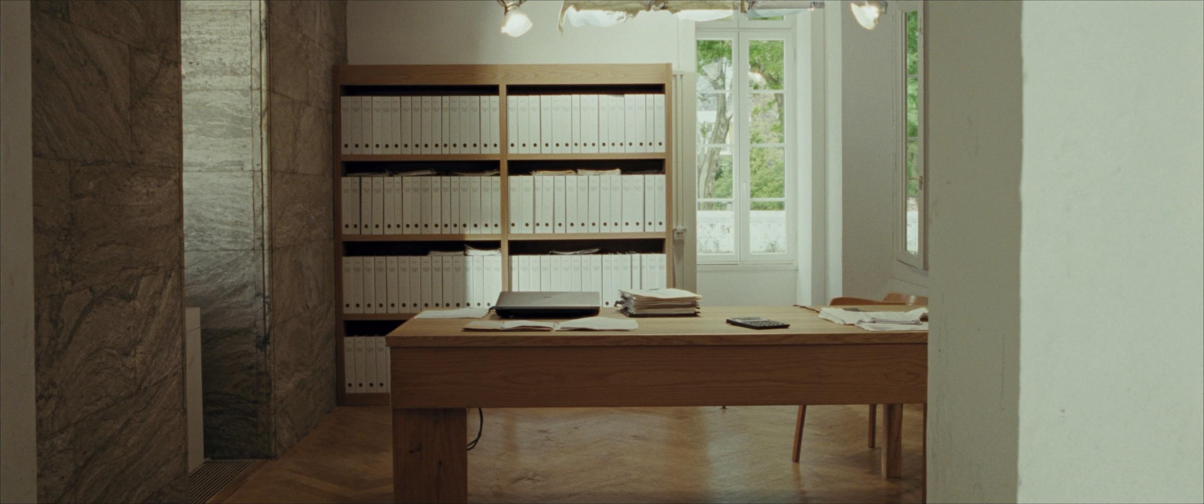 Le bureau d'une fiduciaire : une longue table en chêne, des piles de documents, des classeurs alignés sur des rayonnages en bois, hautes fenêtres.