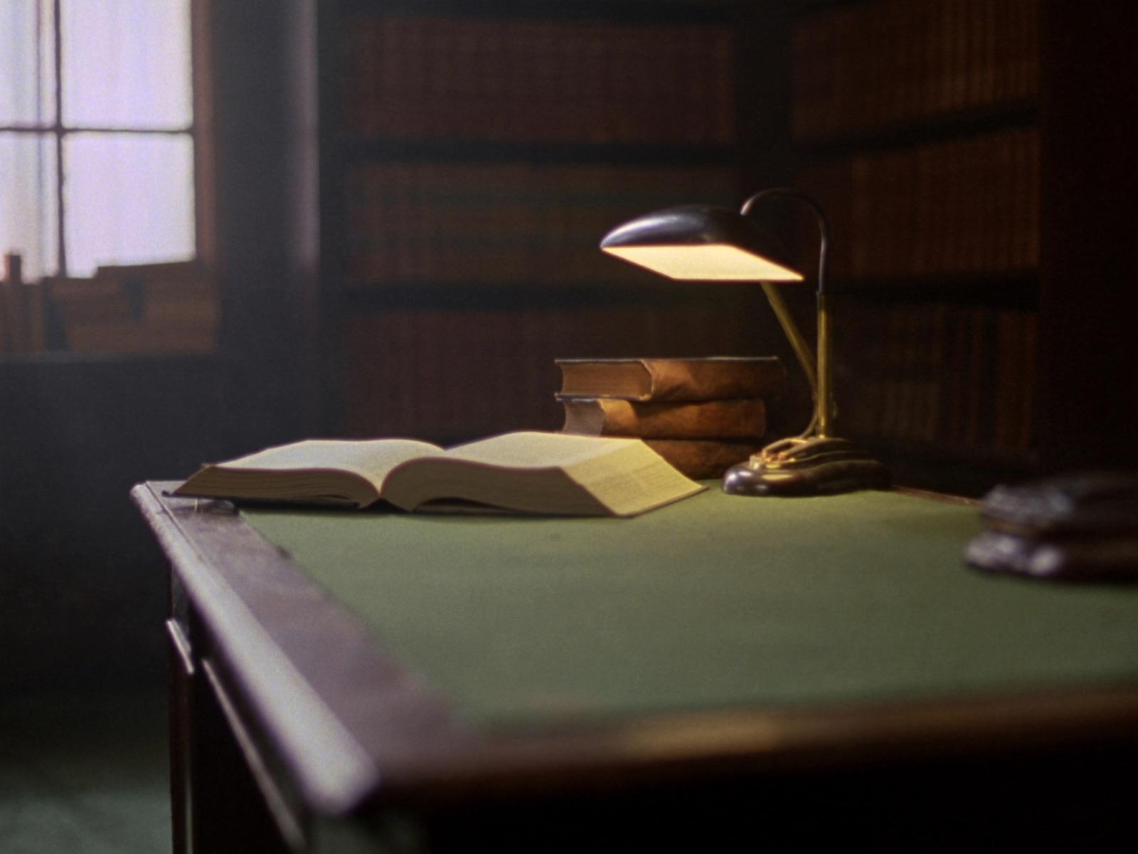 Une table de lecture dans une bibliothèque juridique : un volume ouvert et une pile de codes reliés sous une lampe en laiton.